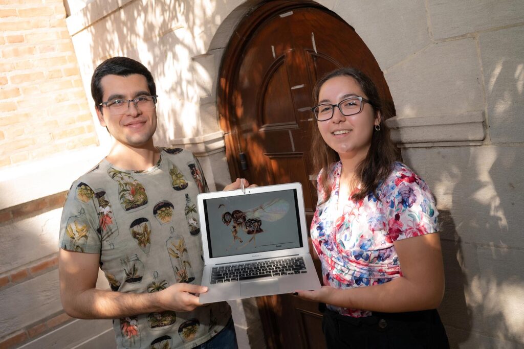 Pedro Brandão-Dias (izquierda) y Camila Vinson afuera de la puerta de Valhalla con una foto de la especie de avispa que no pica que descubrieron cerca. Neuroterus valhalla, que mide solo 1 milímetro de largo, recibió su nombre del pub de estudiantes graduados de Rice porque se descubrió en un árbol en las afueras. Crédito: Jeff Fitlow/Universidad de Rice